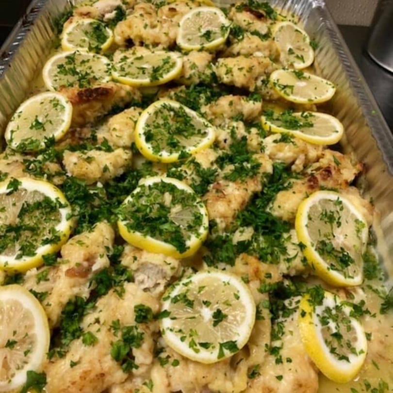 Chicken piccata with lemon slices and fresh parsley in a large catering pan.
