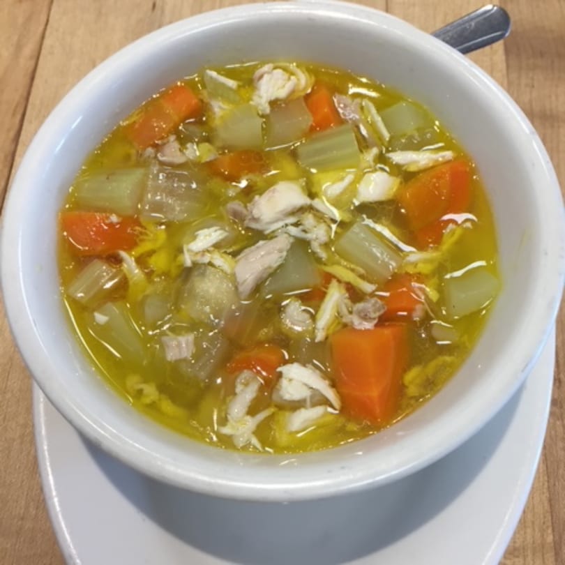 A white bowl of chicken soup with carrots, celery, and shredded chicken on a wooden table.