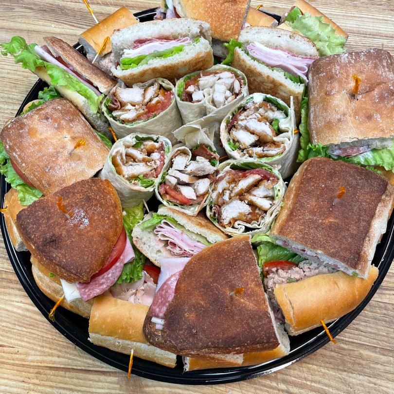 A catering platter from Pizza Village V filled with various sandwiches and wraps, cut in half.